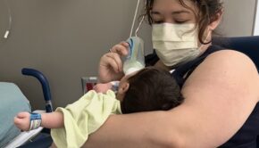A woman wearing a paper mask feeds a bottle to a dark-haired newborn baby in a hospital room