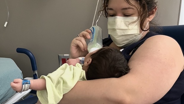 A woman wearing a paper mask feeds a bottle to a dark-haired newborn baby in a hospital room