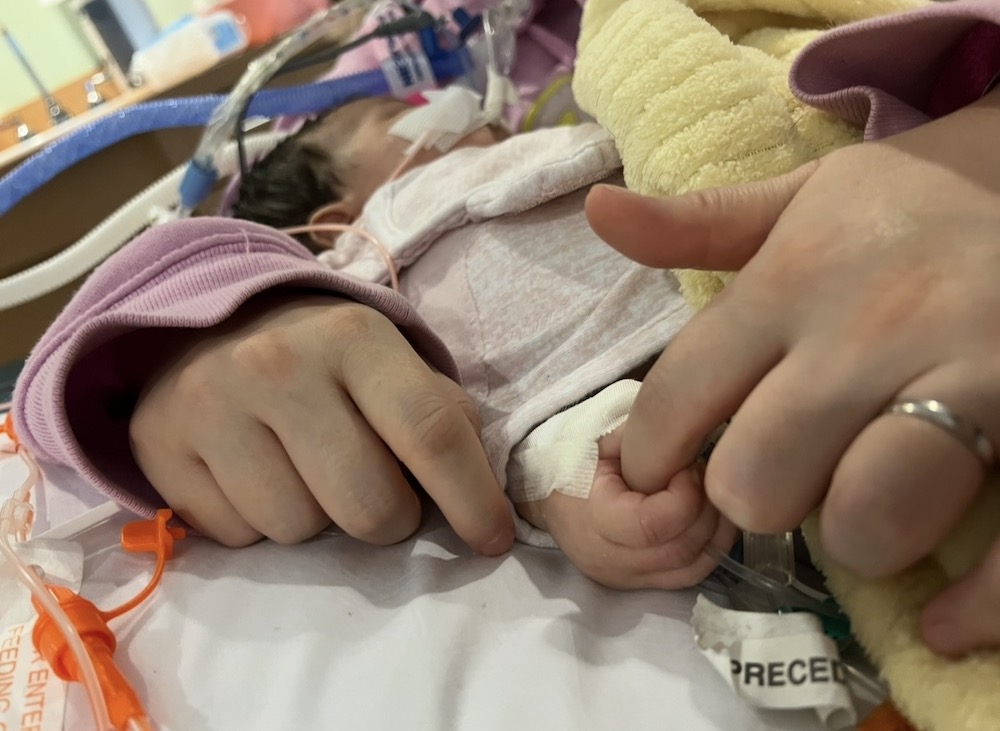 A newborn with tubes taped to her face and hand clutches her mother's finger