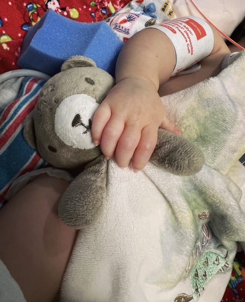 Close-up of a baby's arm with a blood pressure cuff. Her hand rests on a small stuffed bear.