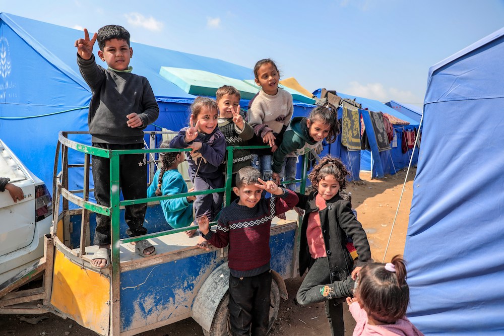 Children in a refugee camp in Gaza ride in a small cart, flashing peace signs