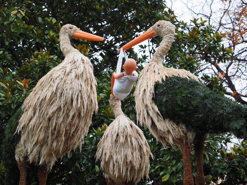 Sculptures of three storks made from natural materials. One holds a plastic human baby in a sling.