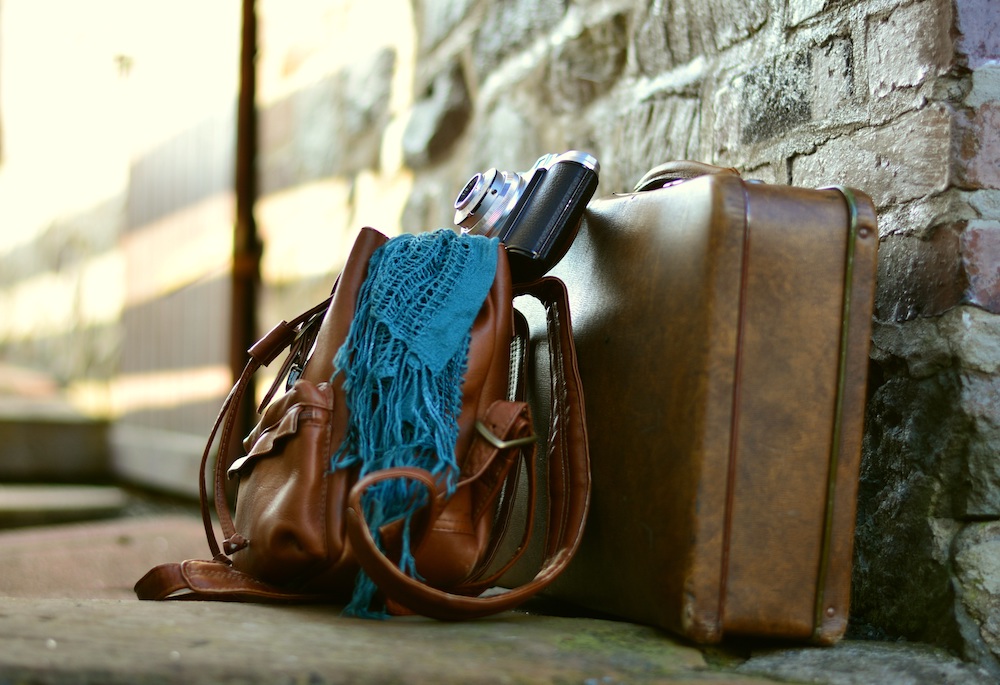 Brown suitcase, brown bag, teal scarf, and camera leaning against a brick wall