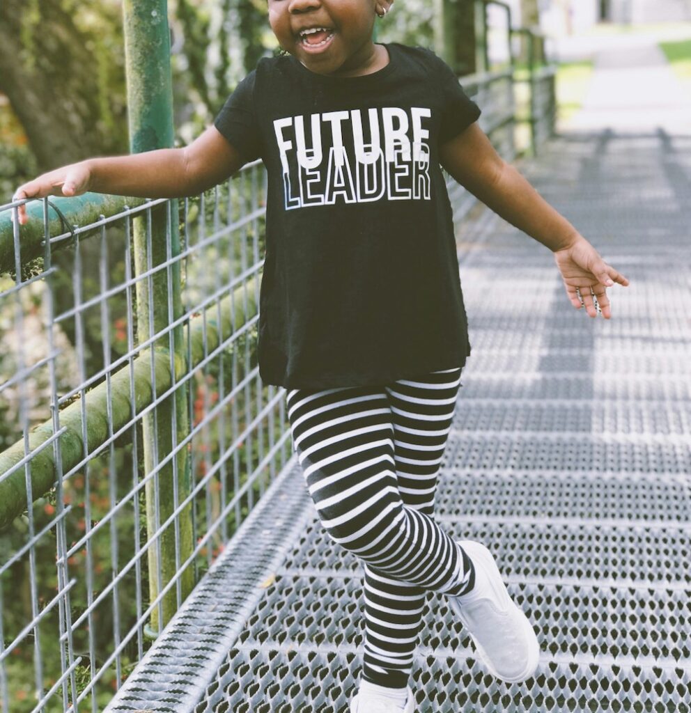 Preschool-age girl with medium brown skin, striped leggings, and a T-shirt that says "Future Leader"