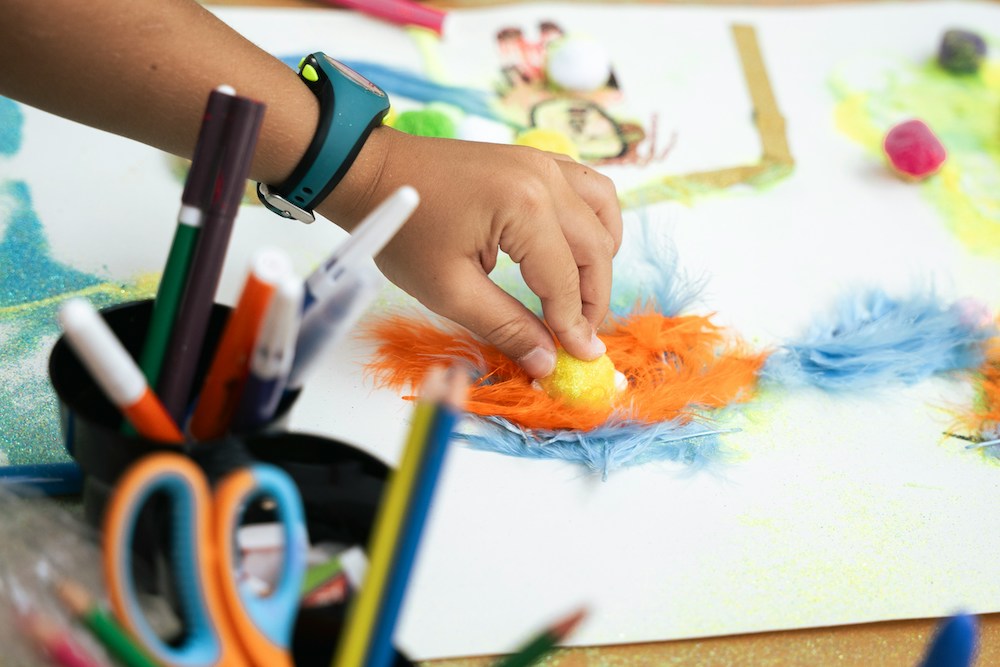 A child's hand places a yellow pom pom on a paper covered in blue and orange feathers