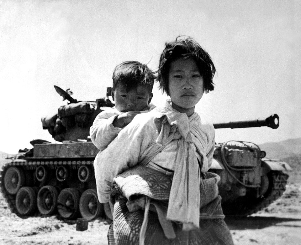 Black and white photo of a Korean girl carrying her toddler brother on her back, standing in front of a tank