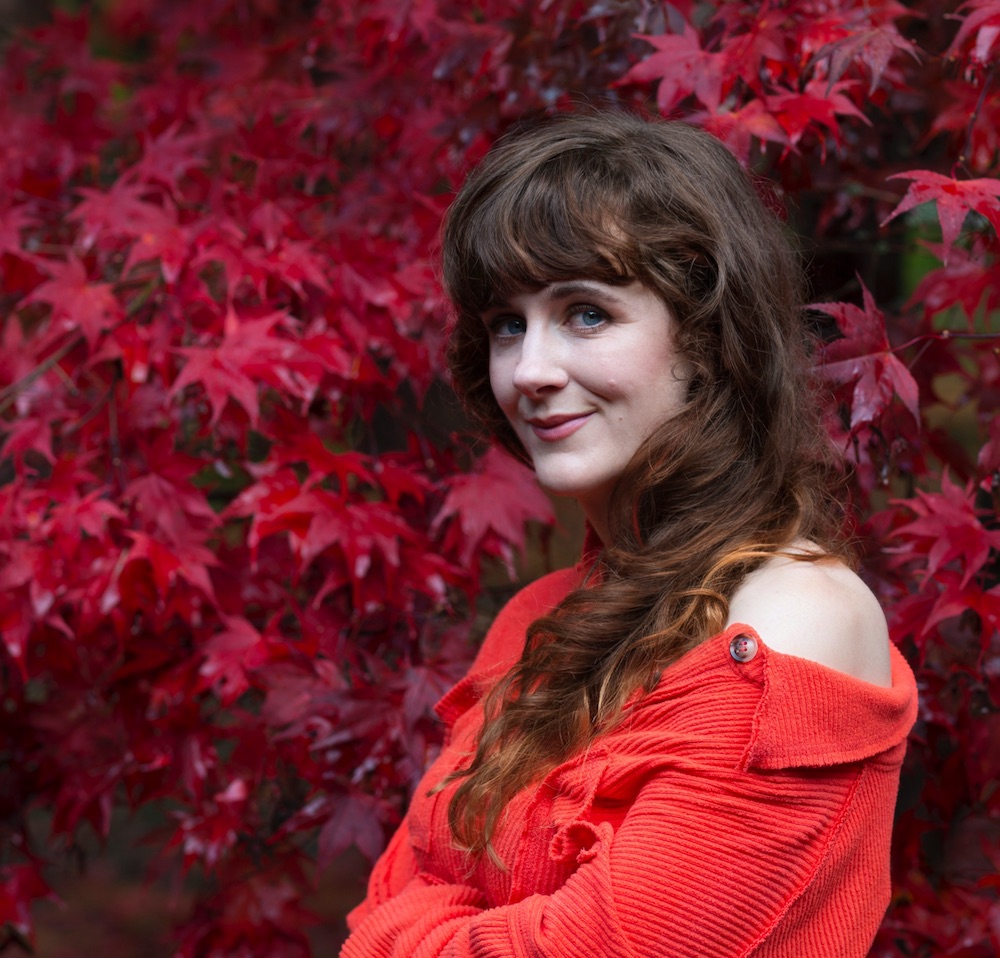 Author photo of Paige Towers, a light skinned woman with wavy auburn hair and an orange top, standing against a background of red leaves.
