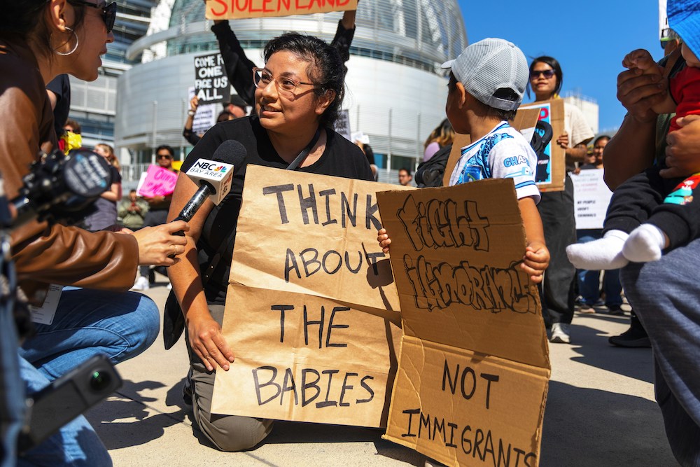 An NBC reporter interviews a woman holding a sign that says "Think about the babies." A small child next to her holds a sign that says "Fight ignorance, not immigrants."