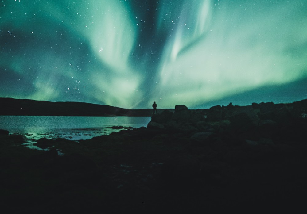 A person standing on black rocks in the distance against a background of blue-green Northern Lights