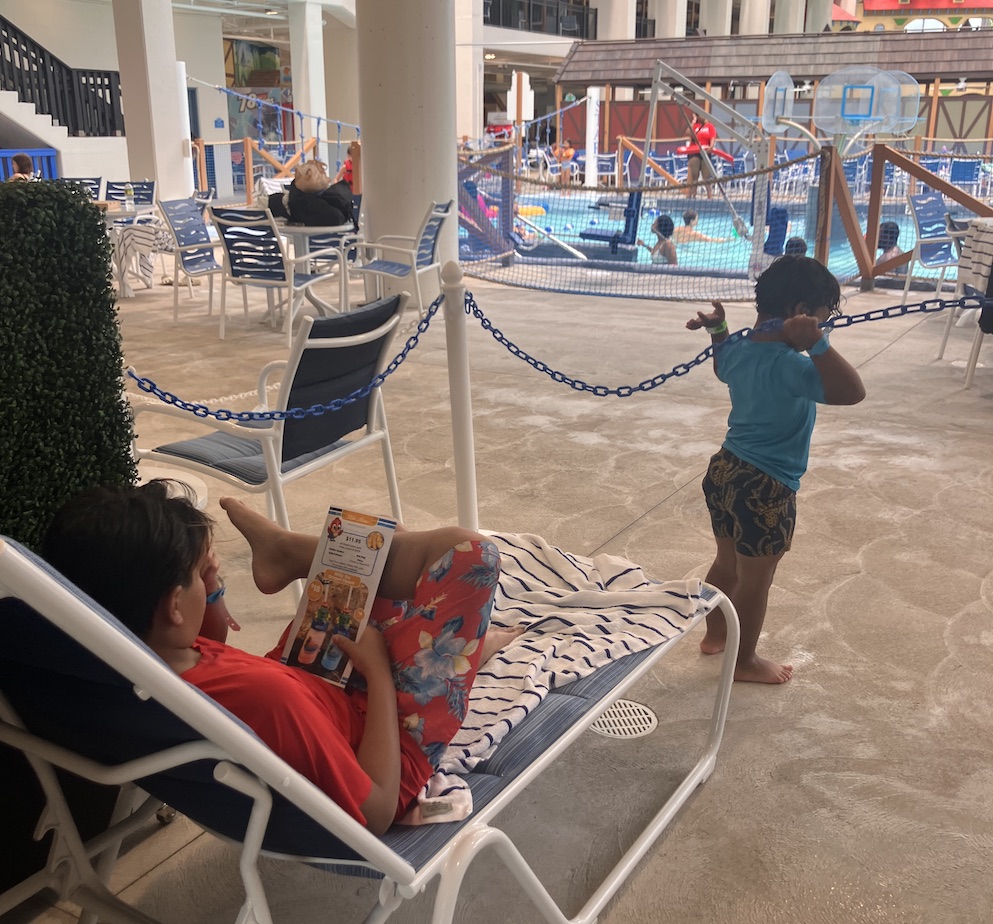 Older child relaxing in a lounge chair while a preschooler plays nearby; a busy pool can be seen in the background