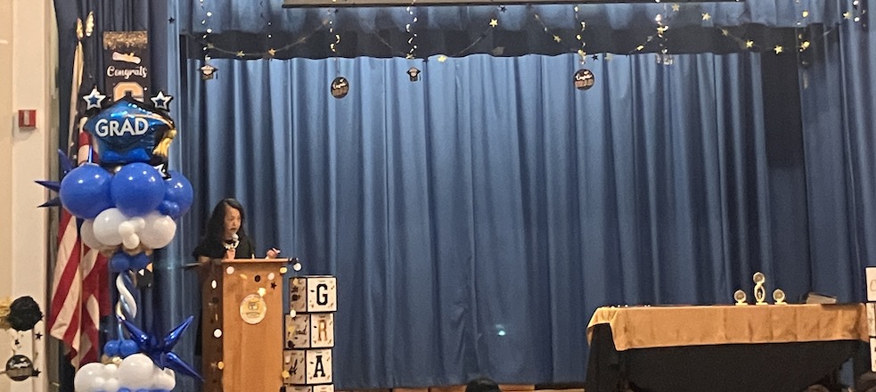 A woman stands at a podium on a stage with blue curtains and graduation decorations
