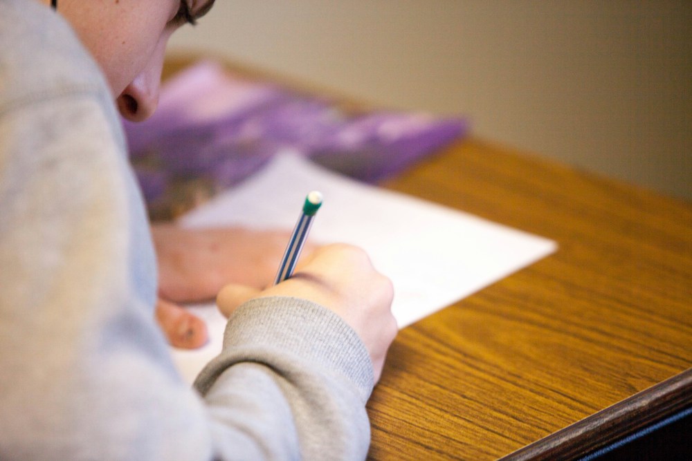 Young person with light skin and gray sweatshirt bent over a paper, writing with a mechanical pencil