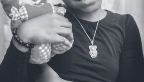 Photo by @ychidy Chidy Young on Unsplash. Black and white photo shows a dark-skinned girl with a teddy bear necklace, clutching a teddy bear