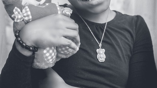 Photo by @ychidy Chidy Young on Unsplash. Black and white photo shows a dark-skinned girl with a teddy bear necklace, clutching a teddy bear