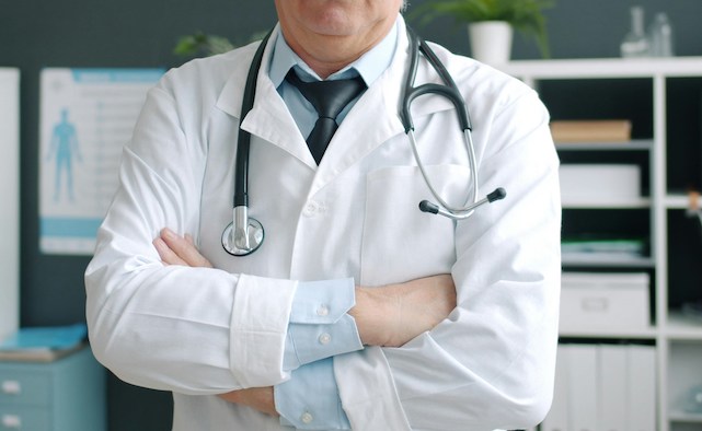 Man in a white lab coat, blue shirt, and tie wears a stethoscope draped over his shoulders and stands with his arms crossed