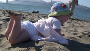 A toddler in a light pink dress and knit hat on her stomach in the sand