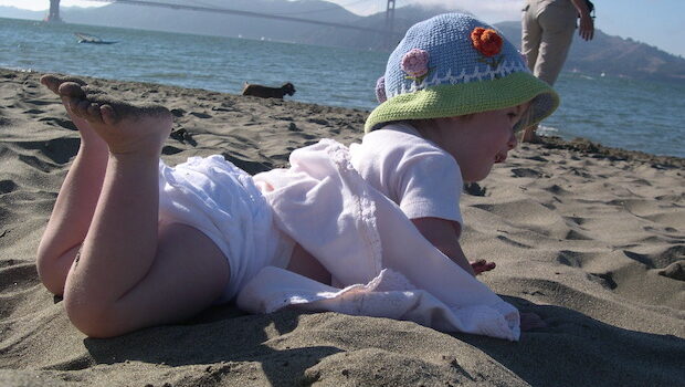 A toddler in a light pink dress and knit hat on her stomach in the sand