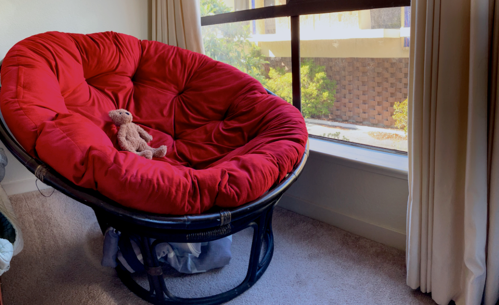 Papasan chair with a large red cushion, holding a teddy bear, next to a large window