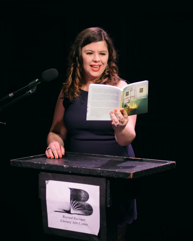 Kelly Grace Thomas, a woman with brown shoulder length hair and red lipstick, reads from a book at a podium