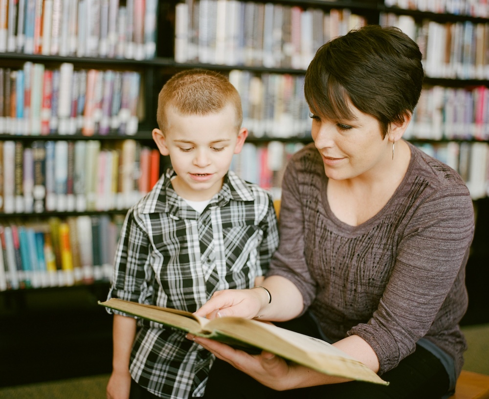 A mother with short brown hair, light skin, and hoop earrings reads to a young boy with short blond hair and a plaid shirt
