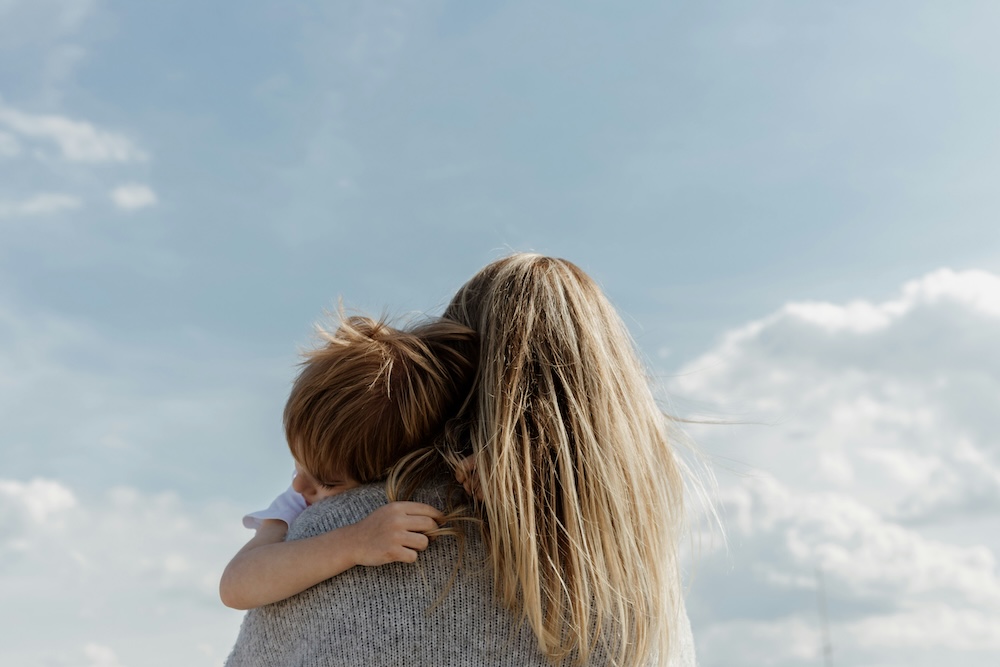 A blonde-haired mother holding a red-headed young child, seen from the back