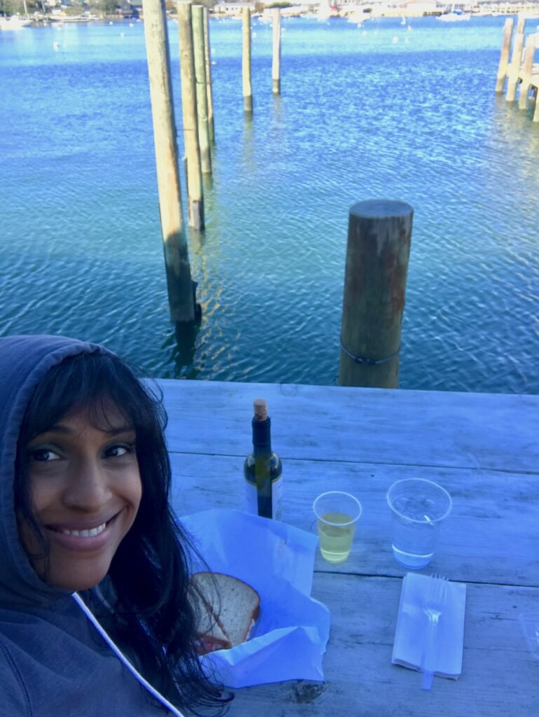 Smiling woman with medium-dark skin and black hair eats a sandwich near the ocean