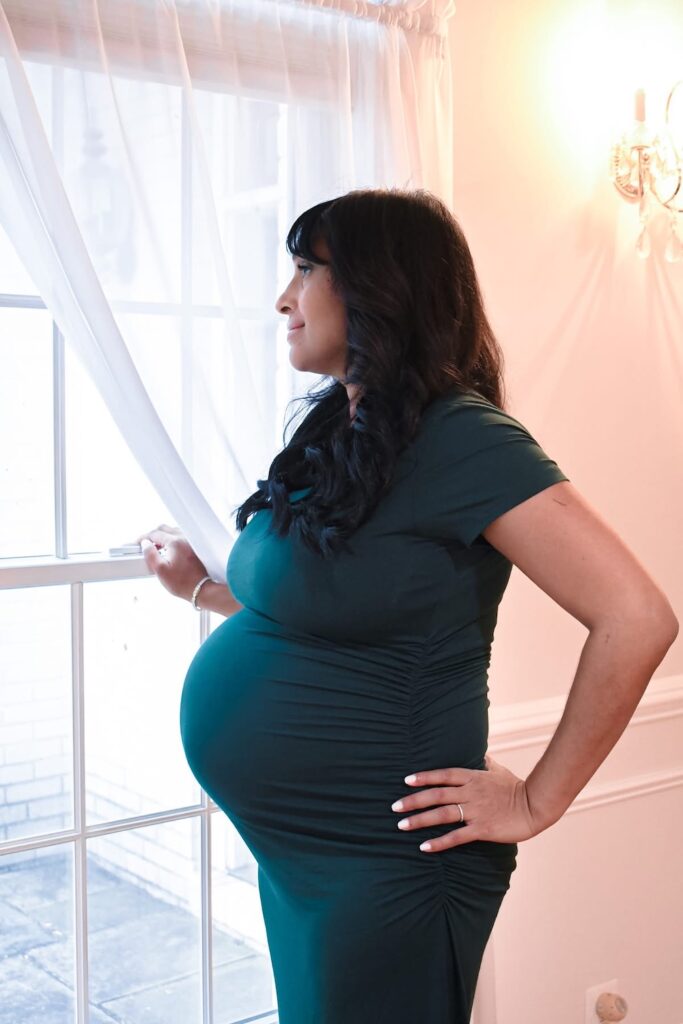 Pregnant woman with brown skin, black hair, and a dark teal dress looks out a window