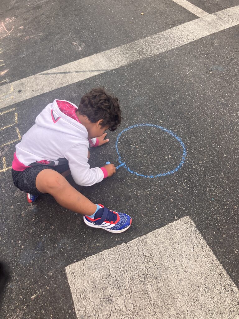 Four-year-old with brown skin and curly brown hair draws a stick figure with blue chalk
