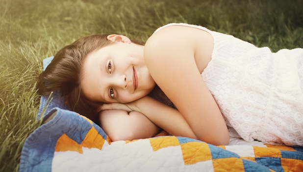 A tween girl with light skin, brown hair, and a slight smile lies on her side on a quilt on the grass. Photo by Jen Theodore.